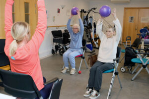 Three women in a rehab center perform seated exercises holding purple balls, guided by an instructor in a coral top.