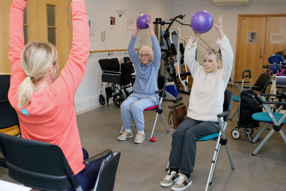 Three women in a rehab center perform seated exercises holding purple balls, guided by an instructor in a coral top.