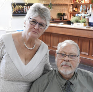 Sharon dressed in a low cut v shaped white dress and older gentleman wearing a olive green shirt with a tie posing inside a warmly lit bar with wooden decor and plants on the counter.