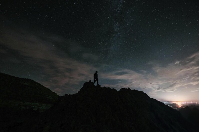 A man stands on a mountain summit under a night sky, illuminated by stars, capturing a moment of reflection and awe.