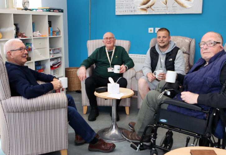 Four men, including one in a wheelchair, sit in a circle holding mugs and chatting in a cosy room with blue walls.