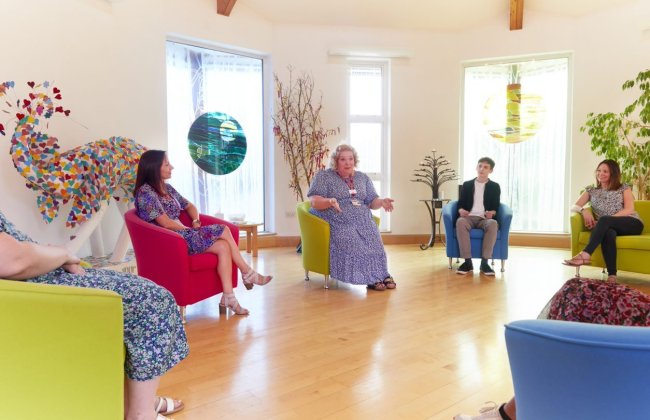 A small group sits together in colourful armchairs inside the Sanctuary, a calm, light-filled space with wooden floors, plants, and stained‑glass-style window art. One person speaks while the others listen, creating a peaceful setting for spiritual care and reflection.
