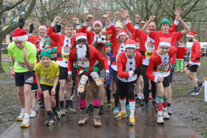 Group of runners dressed in festive holiday attire, including Santa suits and elf hats, posing energetically before a race in a park.