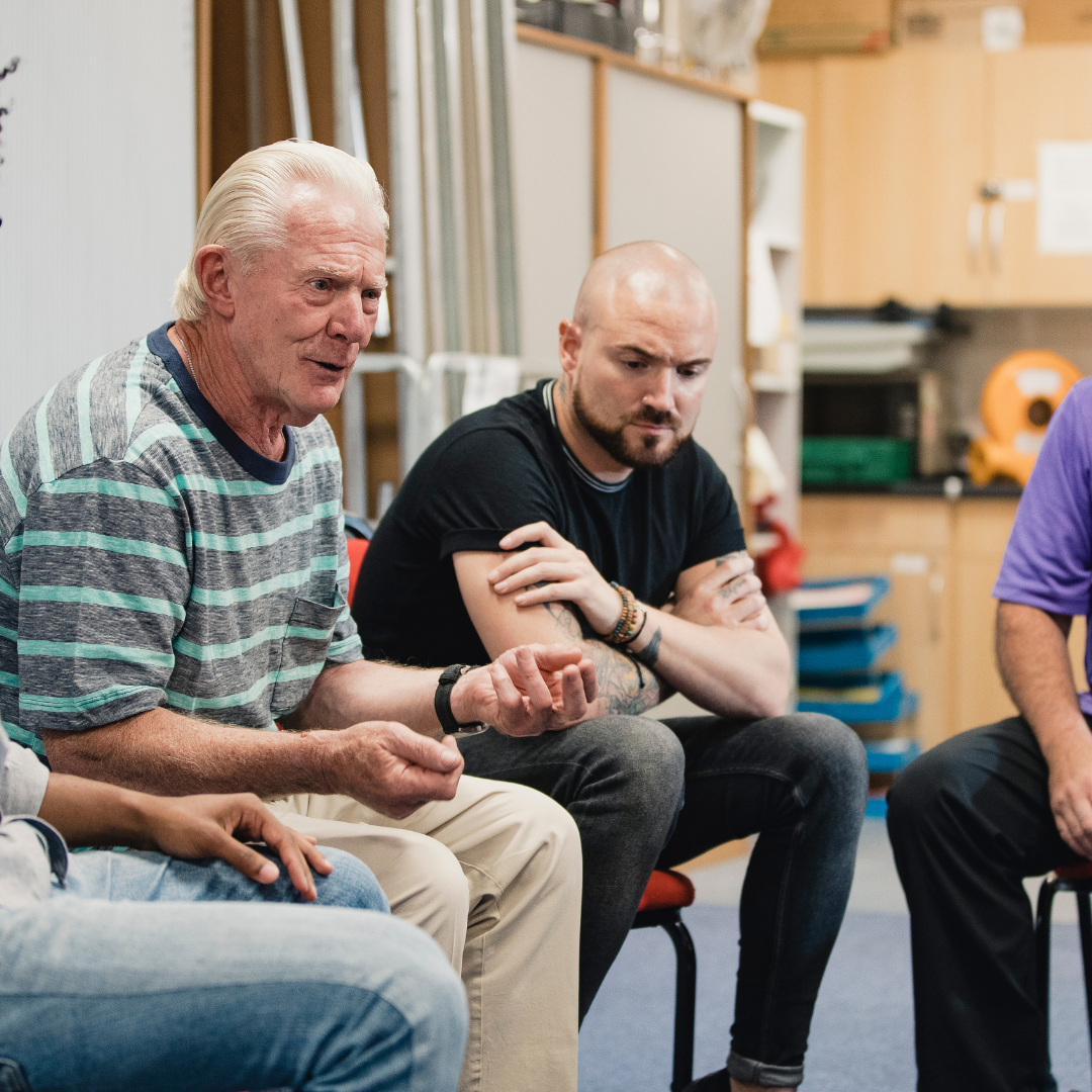 A group of people sit in a circle in a bright indoor room, engaged in conversation during a neuro peer support group. An older man with white hair speaks while others listen attentively, creating a warm, supportive atmosphere.