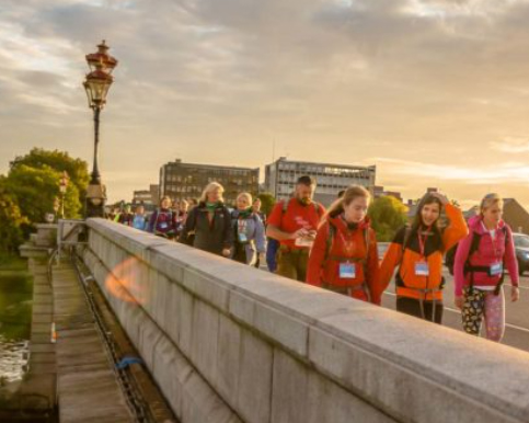 A joyful crowd of people in colourful attire cross a London bridge in the sunshine