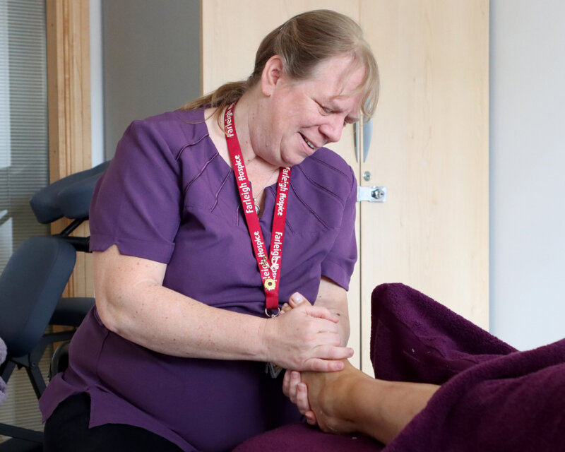 A Farleigh Hospice therapist wearing a purple uniform provides a reflexology foot massage to a patient covered with a purple blanket in a calm treatment room.