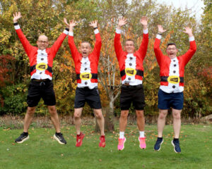Four people wearing festive red Santa-themed shirts and shorts jump with arms raised outdoors amid autumn trees.