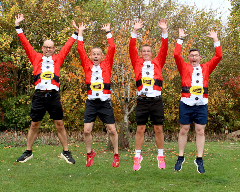 Four people wearing festive red Santa-themed shirts and shorts jump with arms raised outdoors amid autumn trees.