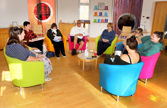 A group of people sit in a circle on brightly coloured chairs in a bright, welcoming room, taking part in a Citizens Advice group session. Notebooks, papers, and cups sit on a small table in the centre as participants talk, listen, and share guidance together.