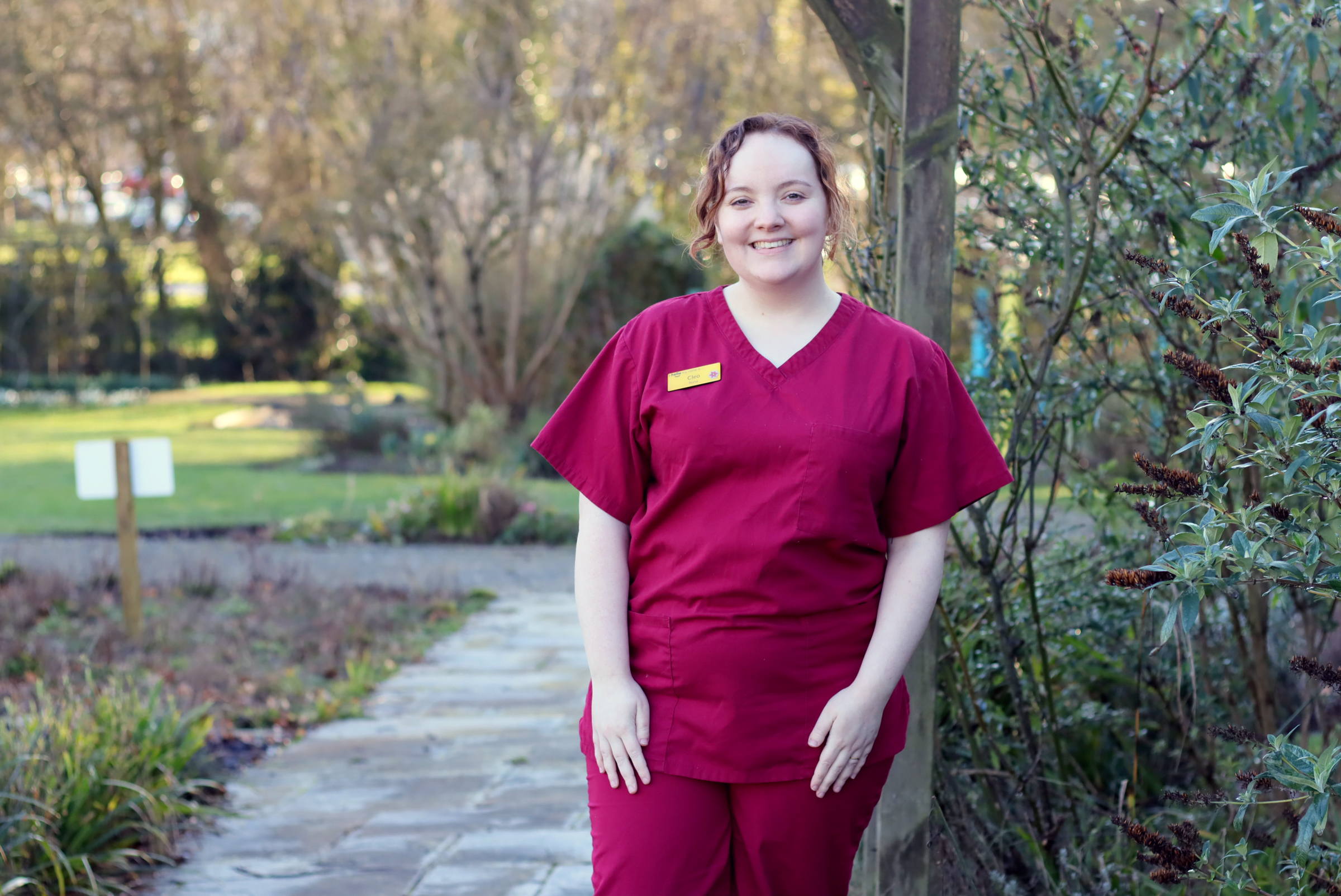 A Farleigh Hospice nurse stands outdoors on a stone path surrounded by greenery, wearing maroon scrubs with a yellow name badge.