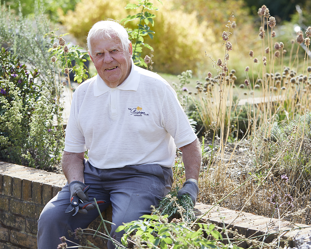 Volunteer gardener Clive sits on the brick wall of a raised bed holding a set of secateurs