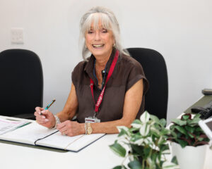 Coralie Taylor, a Farleigh Hospice volunteer, sits at the welcome desk writing in a folder, with a phone and potted plants beside her in a bright, tidy office space.