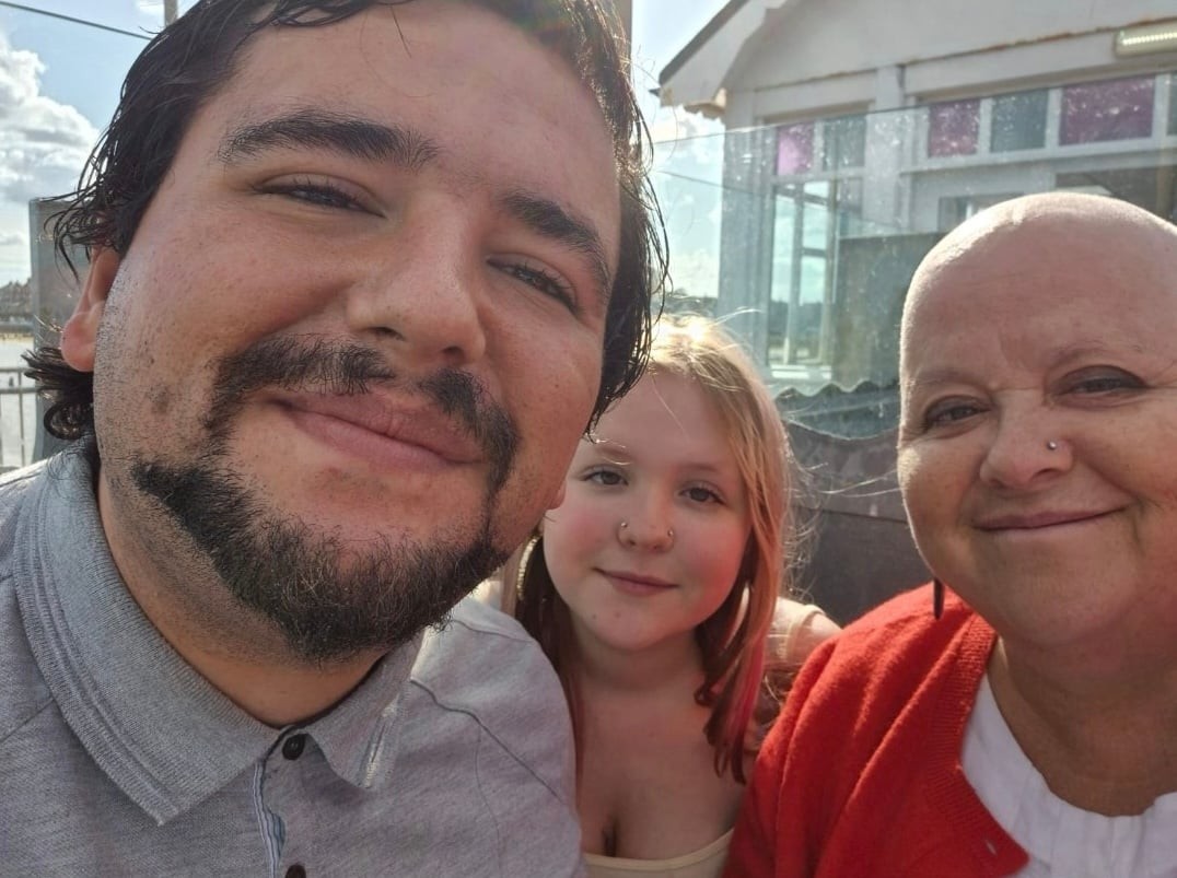 Outdoor selfie of three people smiling in bright sunlight, featuring a young man with dark hair and a short beard in a gray polo shirt, a child with light hair and a nose piercing in the center, and an older woman with a shaved head wearing a red cardigan and nose stud, sitting together on a patio with glass windows and blue sky in the background.