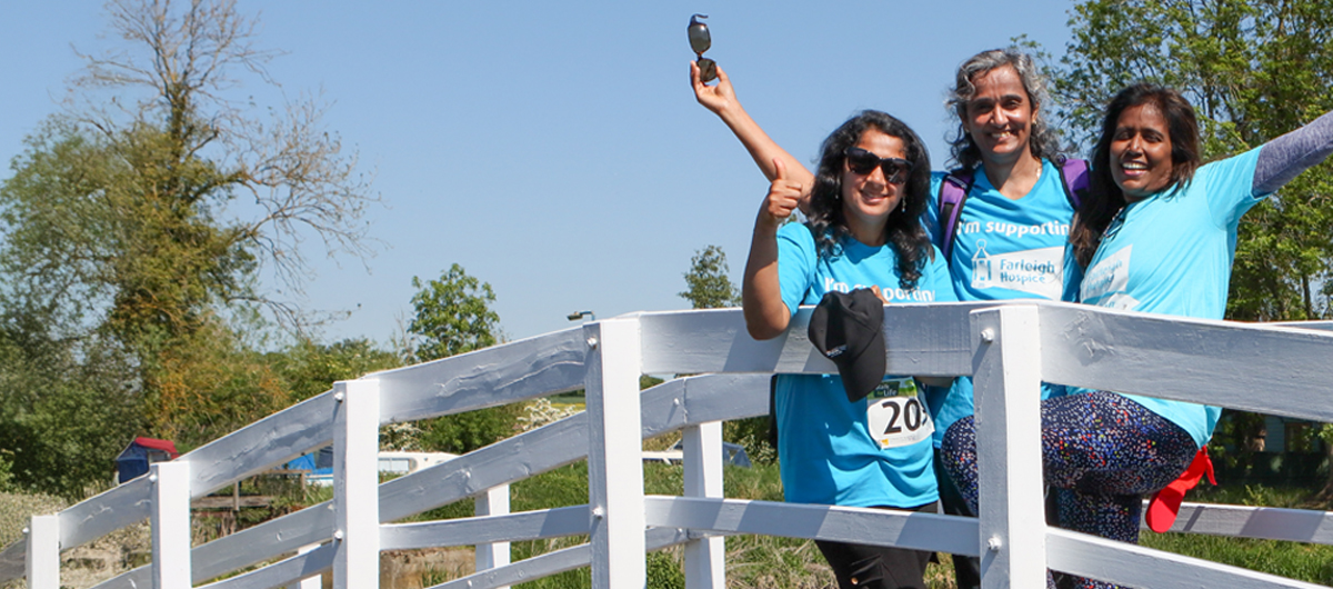 Three participants in blue Farleigh Hospice supporter T‑shirts pause on a bridge during the Walk for Life, with sunny weather and greenery behind them.