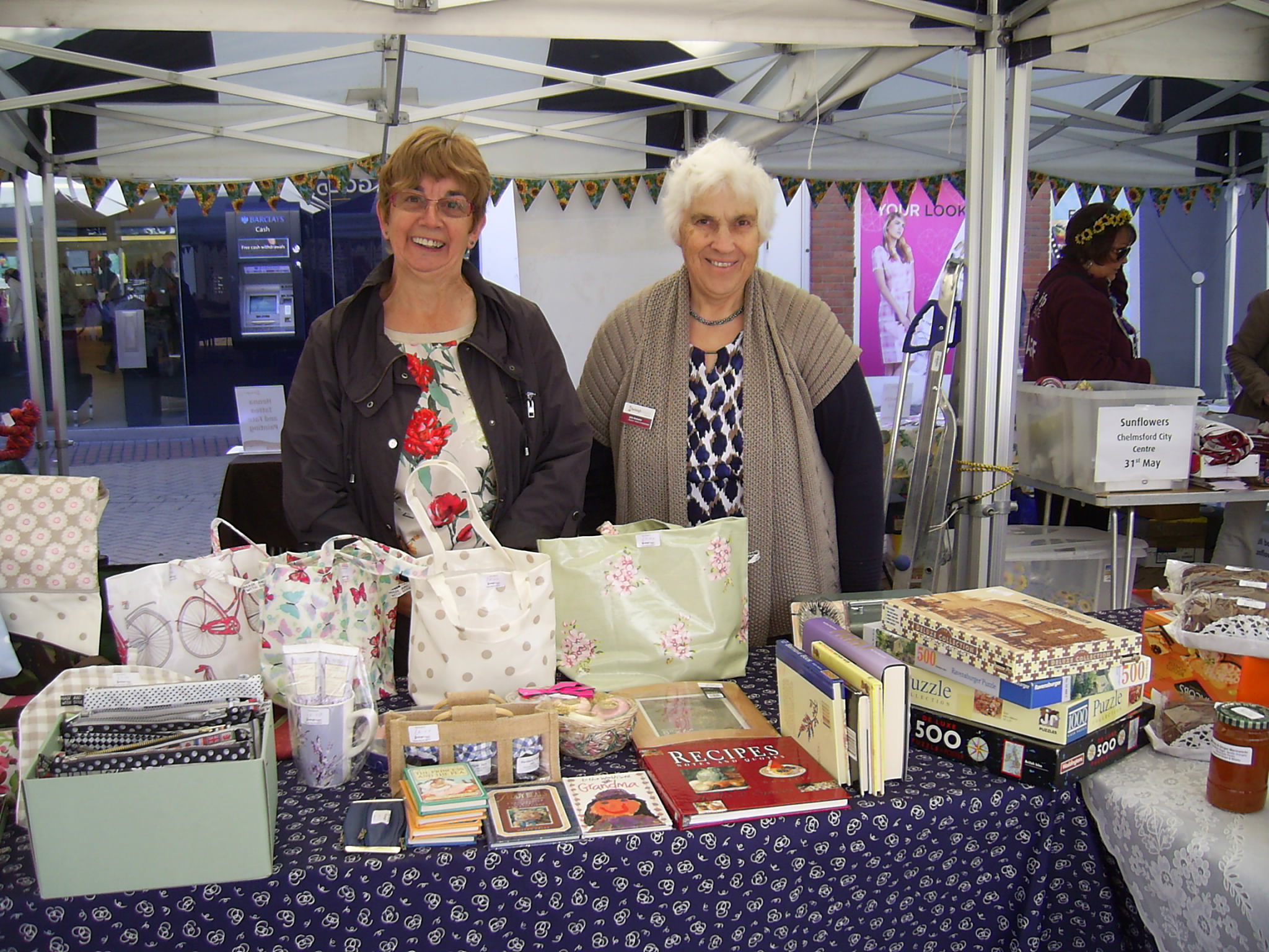 Two female members of the Friends of Farleigh group stand behind a trestle table under a gazebo with craft bags, books and jigsaw puzzles for sale
