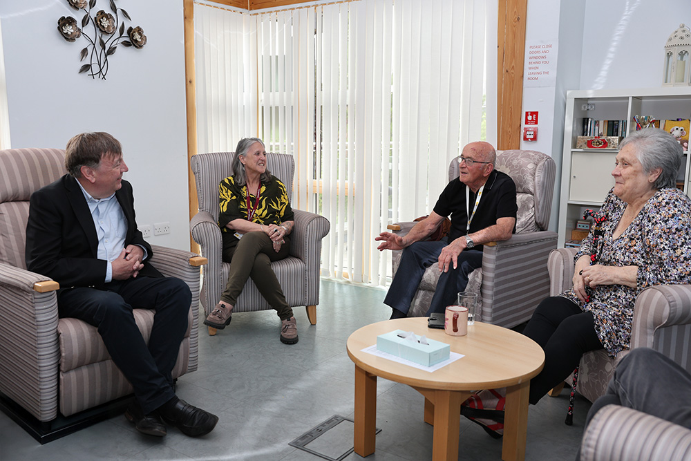 Sir John Whittingdale MP sits in a lounge area speaking to a male Farleigh volunteer and female carer