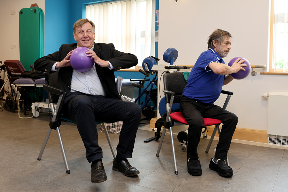 Sir John Whittingdale MP sits in a chair in the gym and raises a lightweight ball as part of a seated exercise class. Another man does the same alongside him.