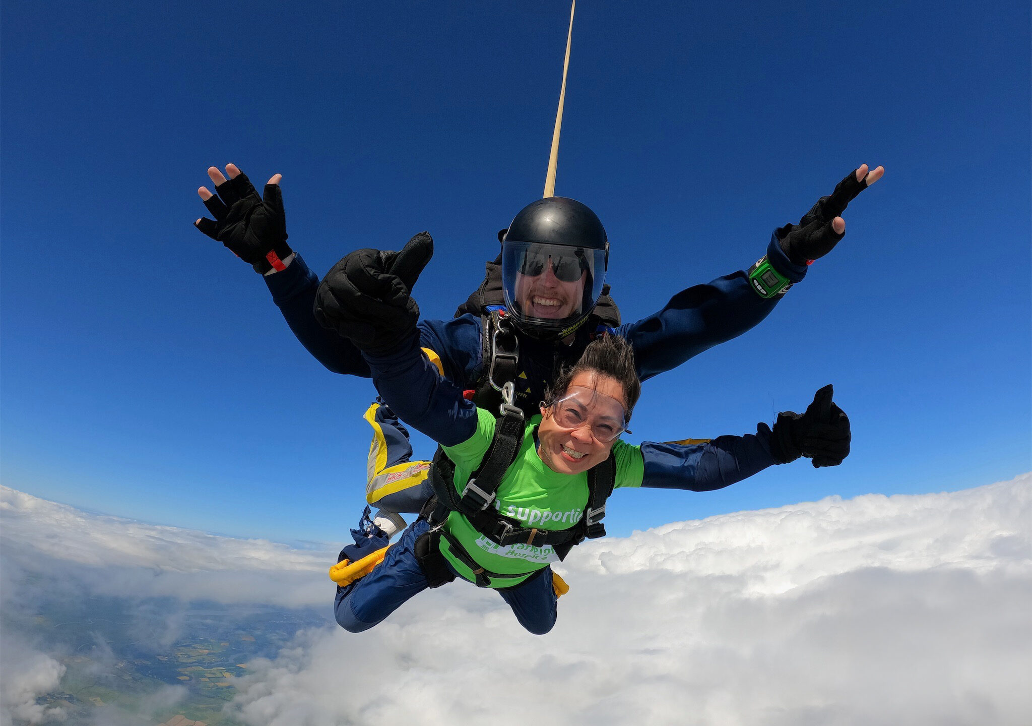 A female skydiver attached to a male instructor in mid flight with blue sky above and clouds below
