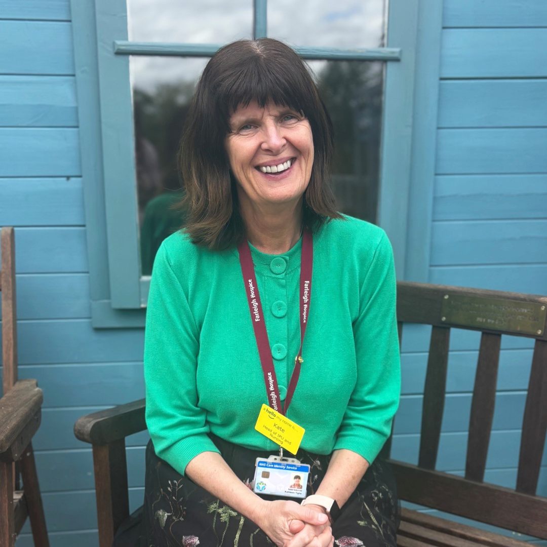 Kate Tighe sits on a wooden bench in front of a teal colour summer house in the garden of Farleigh Hospice