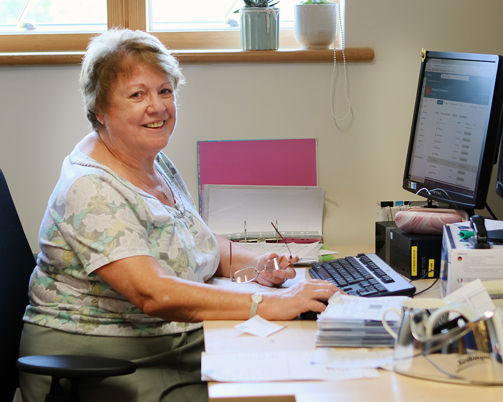 Marilyn, a Farleigh Hospice volunteer, sits at a desk in an office space, working at a computer with paperwork and plants on the windowsill behind her.