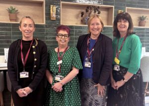 Rachel, Mandy, Ellie and Kate standing together in a staff room, smiling at the camera, each wearing an ID lanyard with a green tiled wall and potted plants behind them.