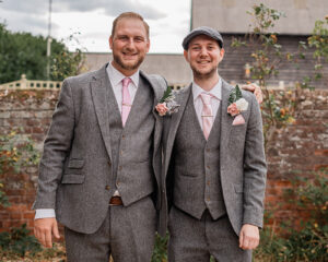 Shane stands on the left of his brother Robson in a walled garden. Both are smartly dressed in grey tweed suits with waistcoats and pink roses in their buttonholes. Shane has his arm around his brother.