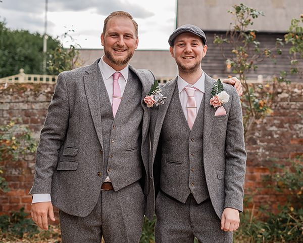 Shane stands on the left of his brother Robson in a walled garden. Both are smartly dressed in grey tweed suits with waistcoats and pink roses in their buttonholes. Shane has his arm around his brother.