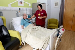 A Farleigh Hospice patient lies in a hospital‑style bed covered with a beige blanket while a Healthcare Assistant in a maroon Farleigh uniform with a yellow name badge stands beside them, speaking gently and checking on their comfort. The room is calm and homely, with medical equipment, a bedside cabinet, two chairs, framed artwork on the wall and a wooden door.