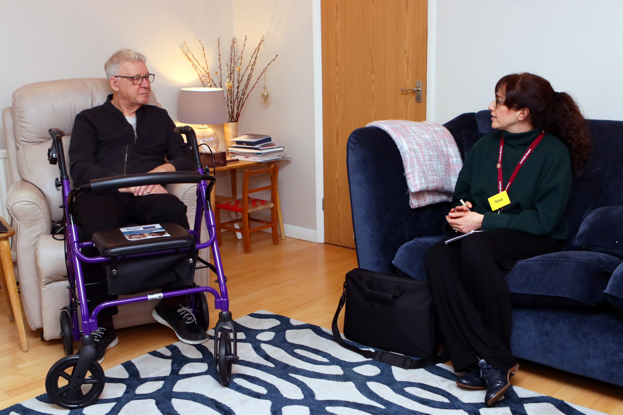 Tessa Rose, Farleigh Hospice’s Motor Neurone Disease Coordinator, sits with patient Richard in his home, talking together in a comfortable living‑room setting with a walker positioned nearby.