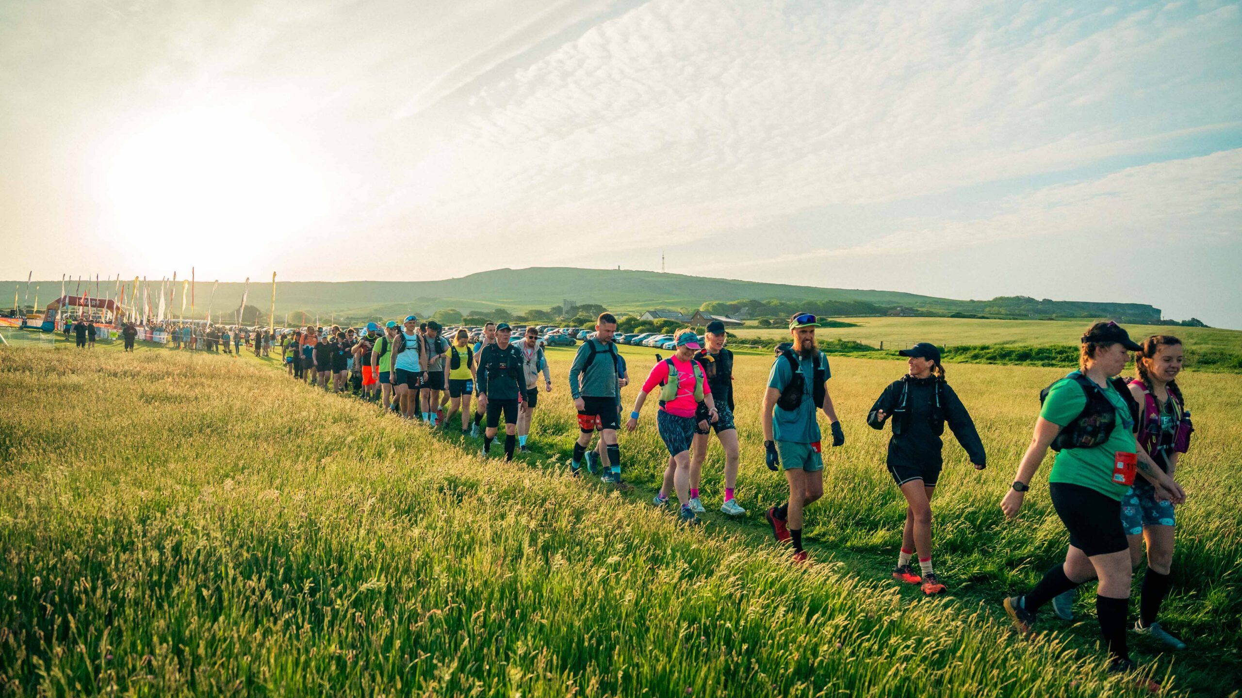 A cheerful group of hikers in colorful outdoor gear walks through a sunlit meadow, with rolling green hills and a bright sky behind them.