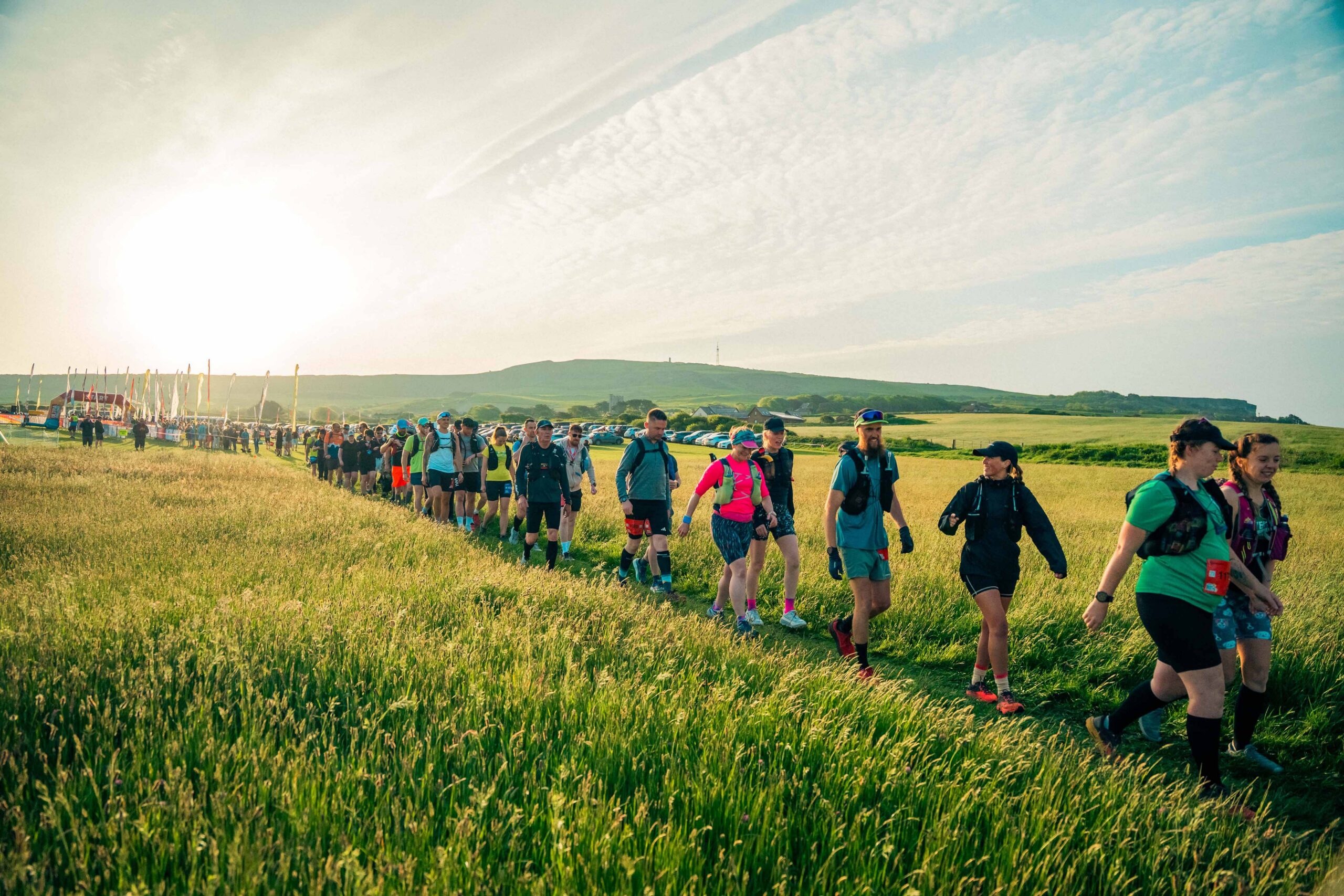 A cheerful group of hikers in colorful outdoor gear walks through a sunlit meadow, with rolling green hills and a bright sky behind them.