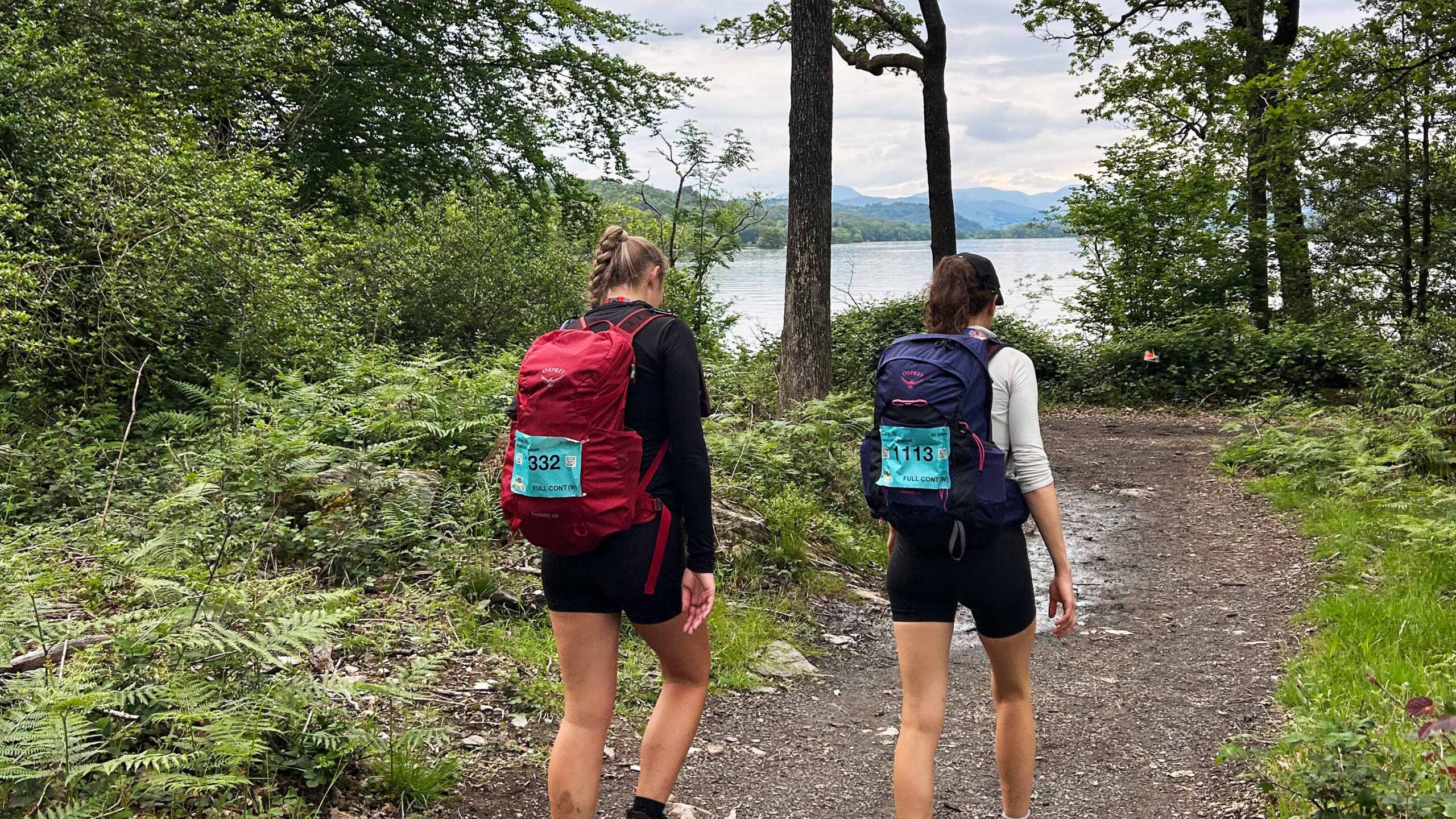 Two female hikers with numbered backpacks walk a wooded trail toward a scenic lake and mountains, creating a peaceful outdoor adventure scene.