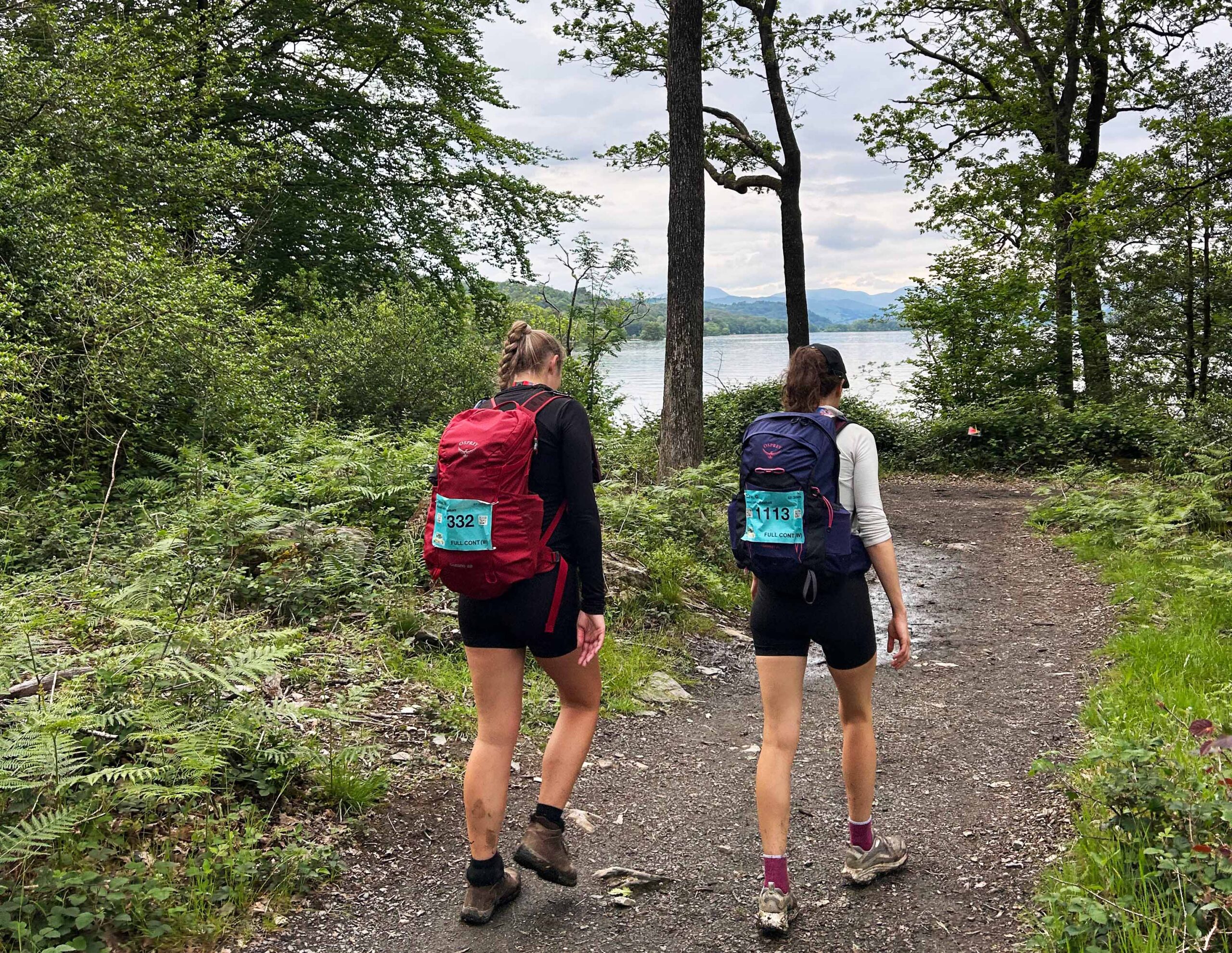 Two female hikers with numbered backpacks walk a wooded trail toward a scenic lake and mountains, creating a peaceful outdoor adventure scene.