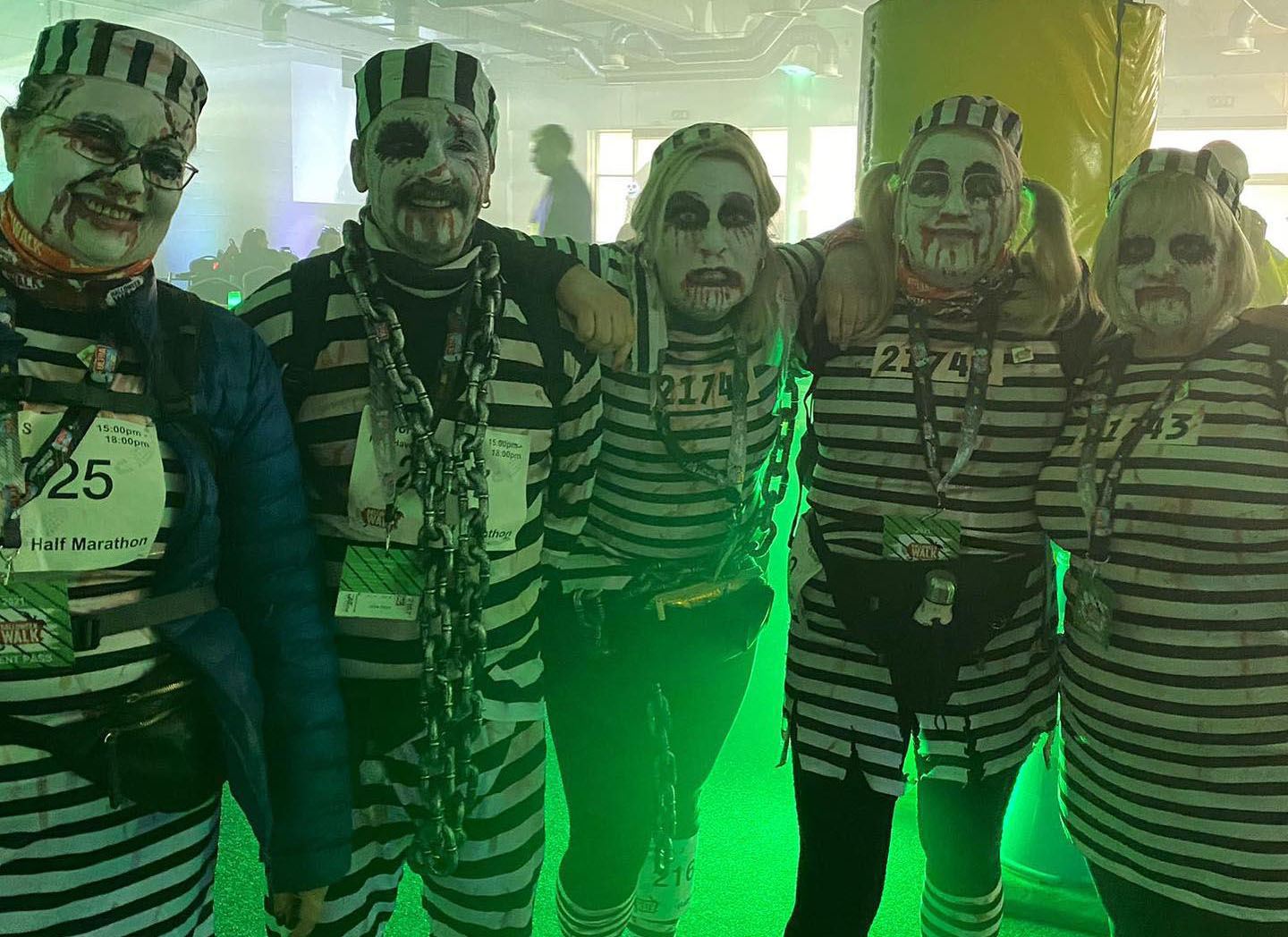 Five people in striped prisoner costumes and skull makeup pose at a walking event, smiling under eerie green lighting in a playful spooky scene.