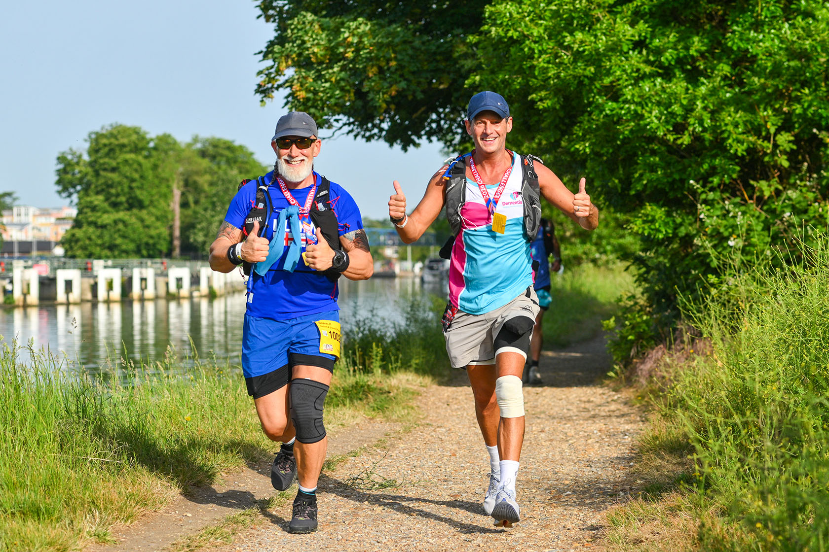 Two smiling male runners in colorful athletic gear jog along a sunny riverside trail, giving thumbs-up; lush greenery and calm water surround them.