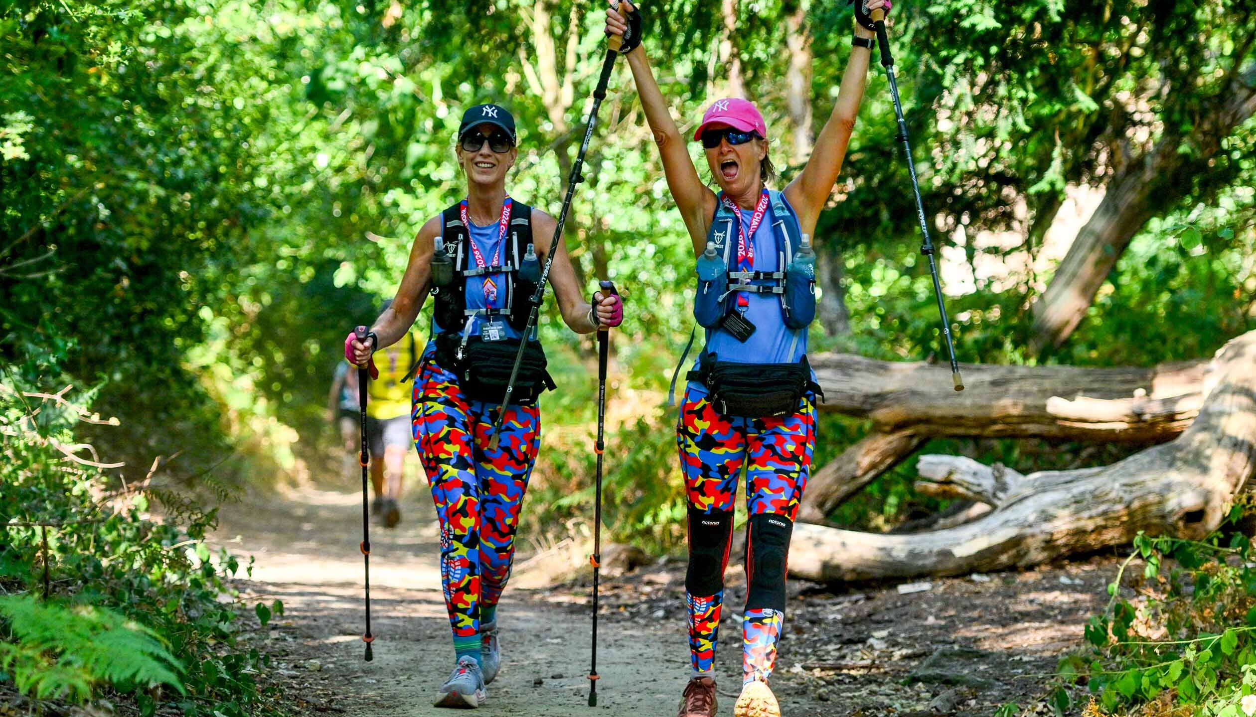 Two cheerful women hikers in colorful clothing celebrate on a forest trail, raising trekking poles amid sunlit trees and fallen logs.