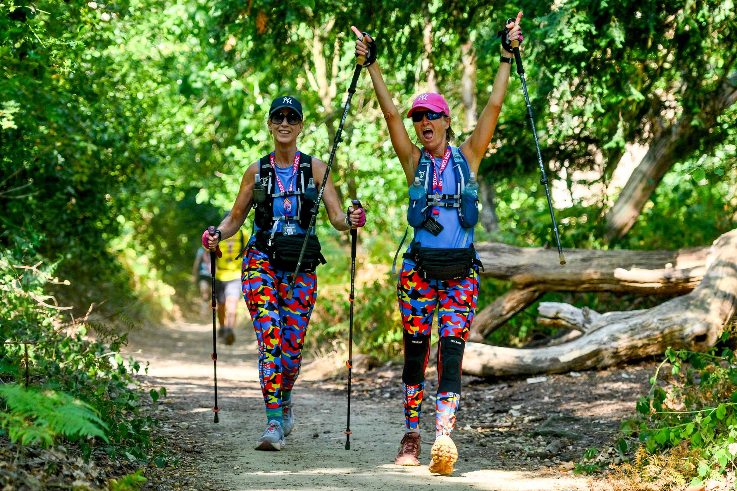 Two cheerful women hikers in colorful clothing celebrate on a forest trail, raising trekking poles amid sunlit trees and fallen logs.
