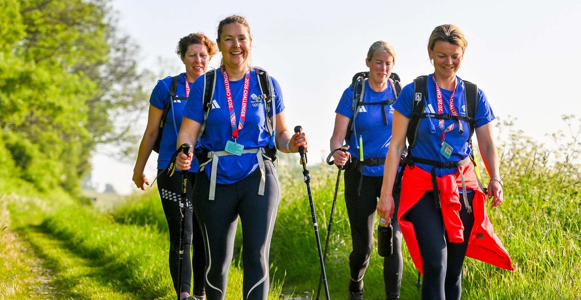 Four cheerful hikers in blue shirts walk together along a sunny green countryside trail, carrying backpacks and trekking poles, conveying camaraderie.