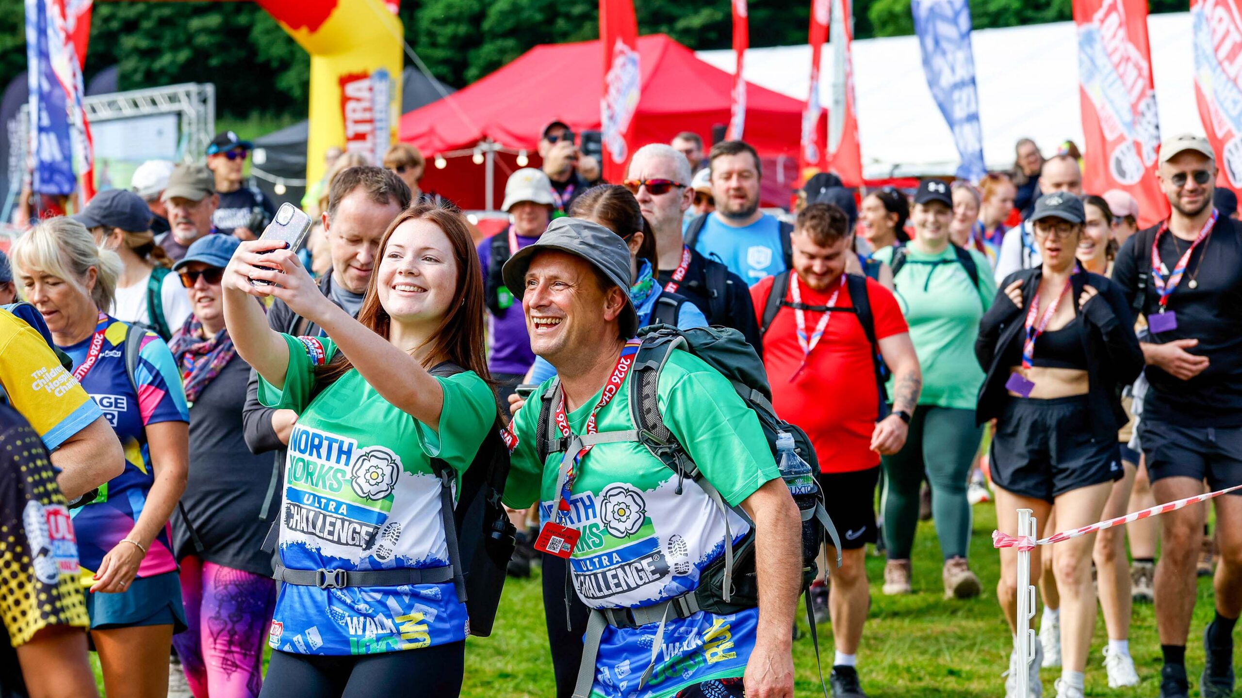 Smiling race participants take a selfie at an outdoor challenge event, surrounded by colorful team shirts, flags, tents, and cheering walkers.