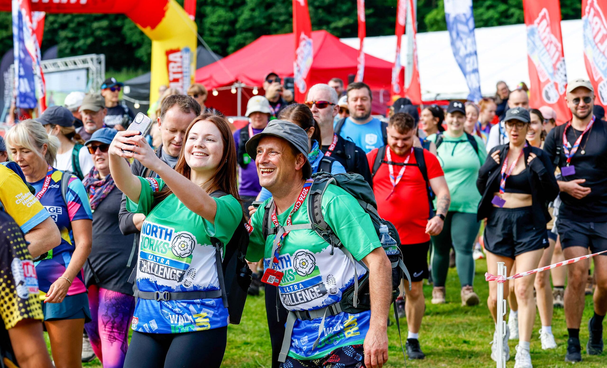 Smiling race participants take a selfie at an outdoor challenge event, surrounded by colorful team shirts, flags, tents, and cheering walkers.