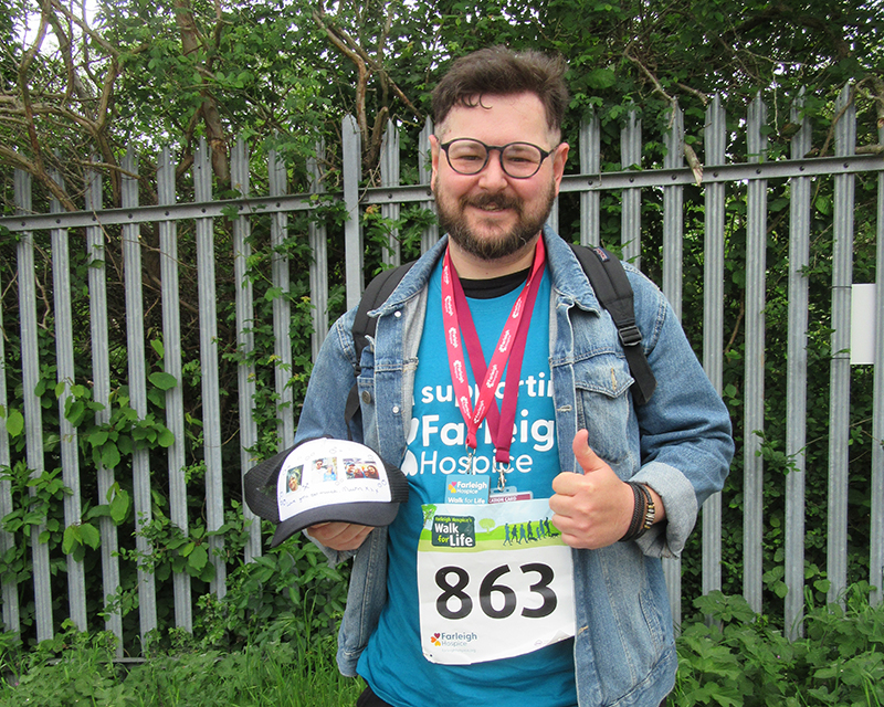 Samuel Brewster stands in the car park at the end of the Walk for Life event with one thumb up holding a baseball cap that features photos of his mother