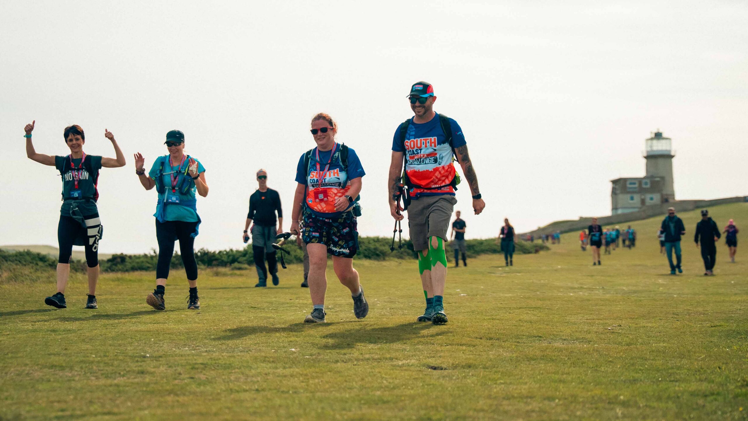 Hikers in colorful athletic clothing walk across a grassy coastal field, smiling and waving, with a lighthouse and distant walkers behind them.