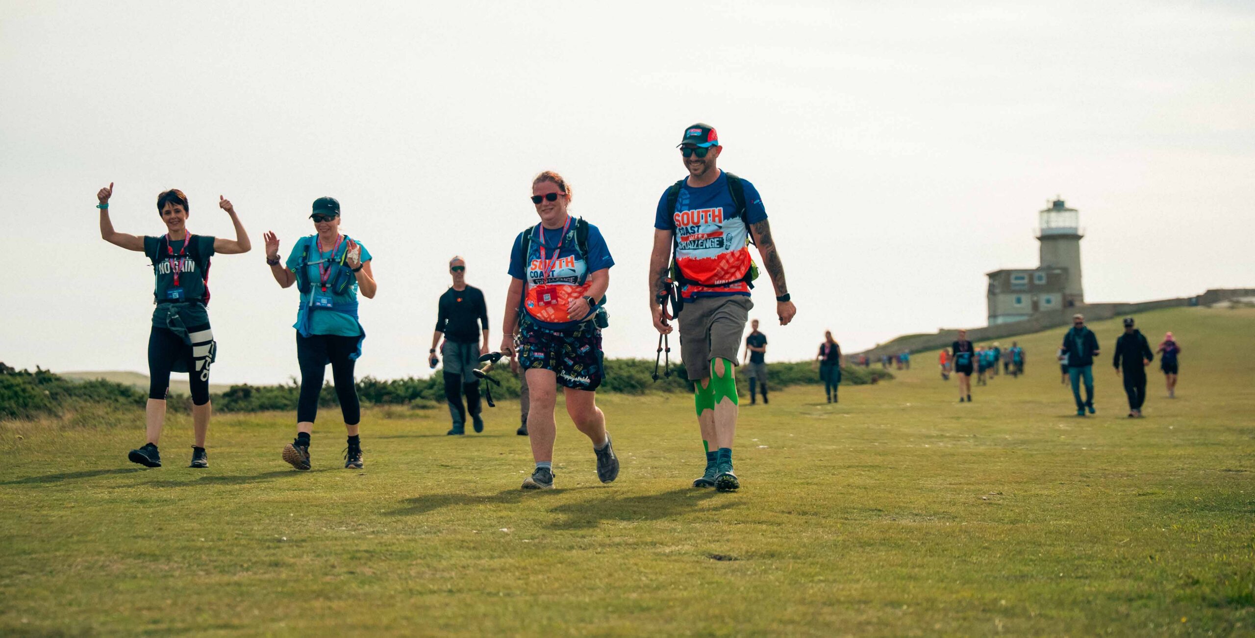 Hikers in colorful athletic clothing walk across a grassy coastal field, smiling and waving, with a lighthouse and distant walkers behind them.
