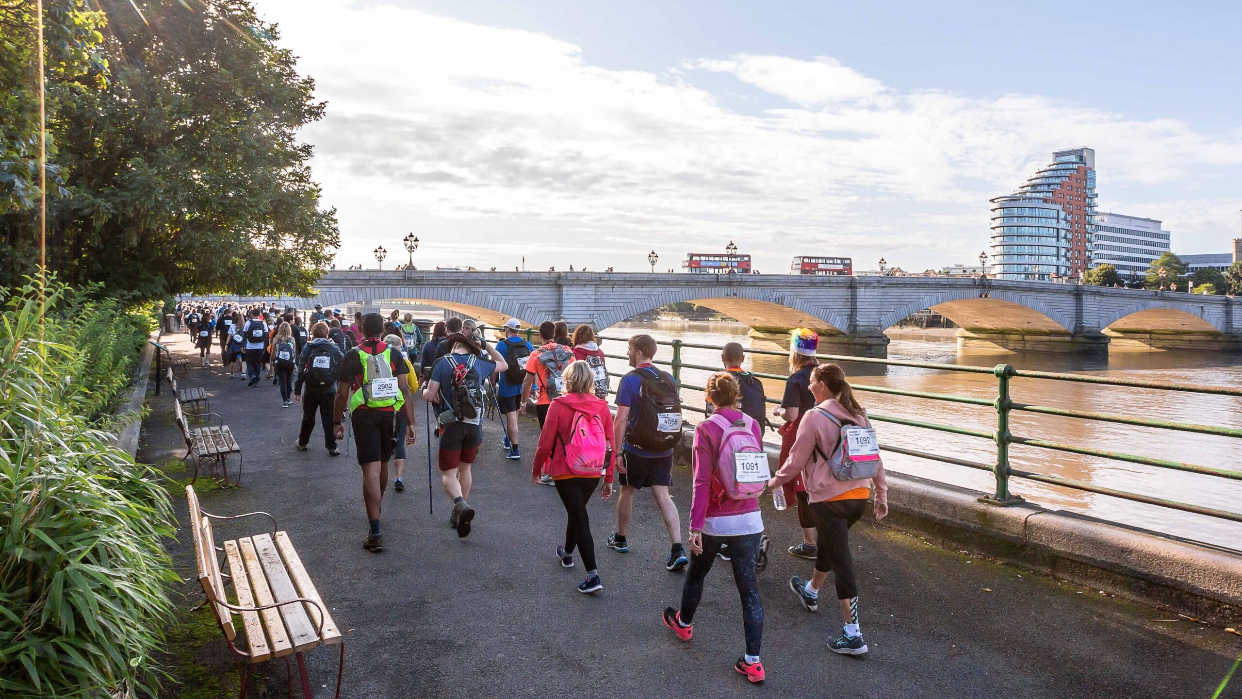Walkers in colourful charity race shirts follow a riverside path beneath sunny skies, with a bridge, river, and modern city buildings behind.