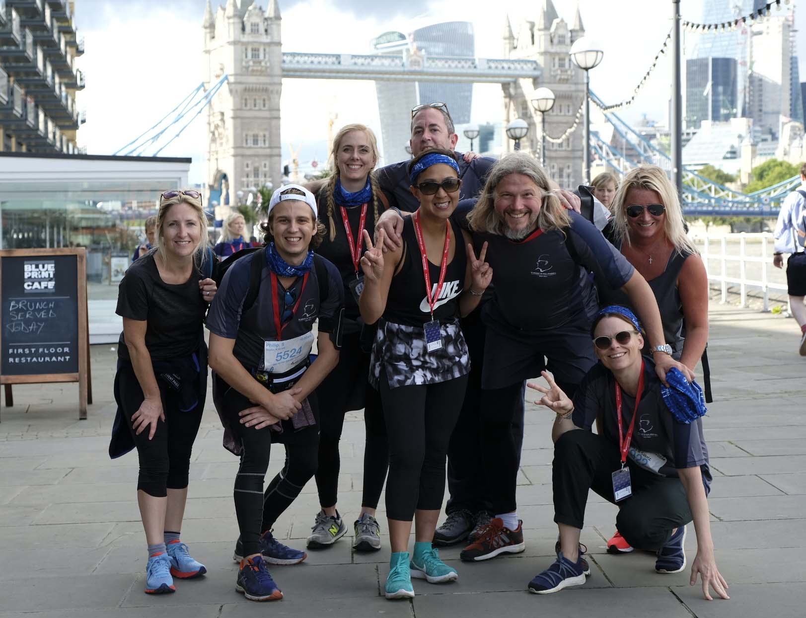 Smiling group of eight runners pose on London’s riverside promenade, with Tower Bridge and skyscrapers behind them on a bright day