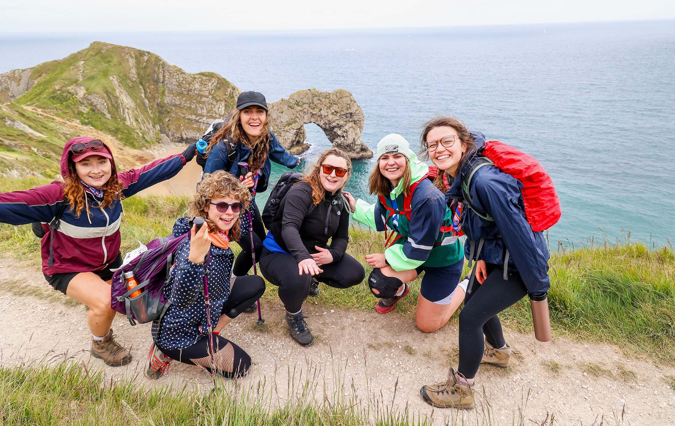 Six cheerful hikers pose on a grassy coastal cliff trail in colorful outdoor gear and backpacks, with the blue ocean and rocky headland behind them.
