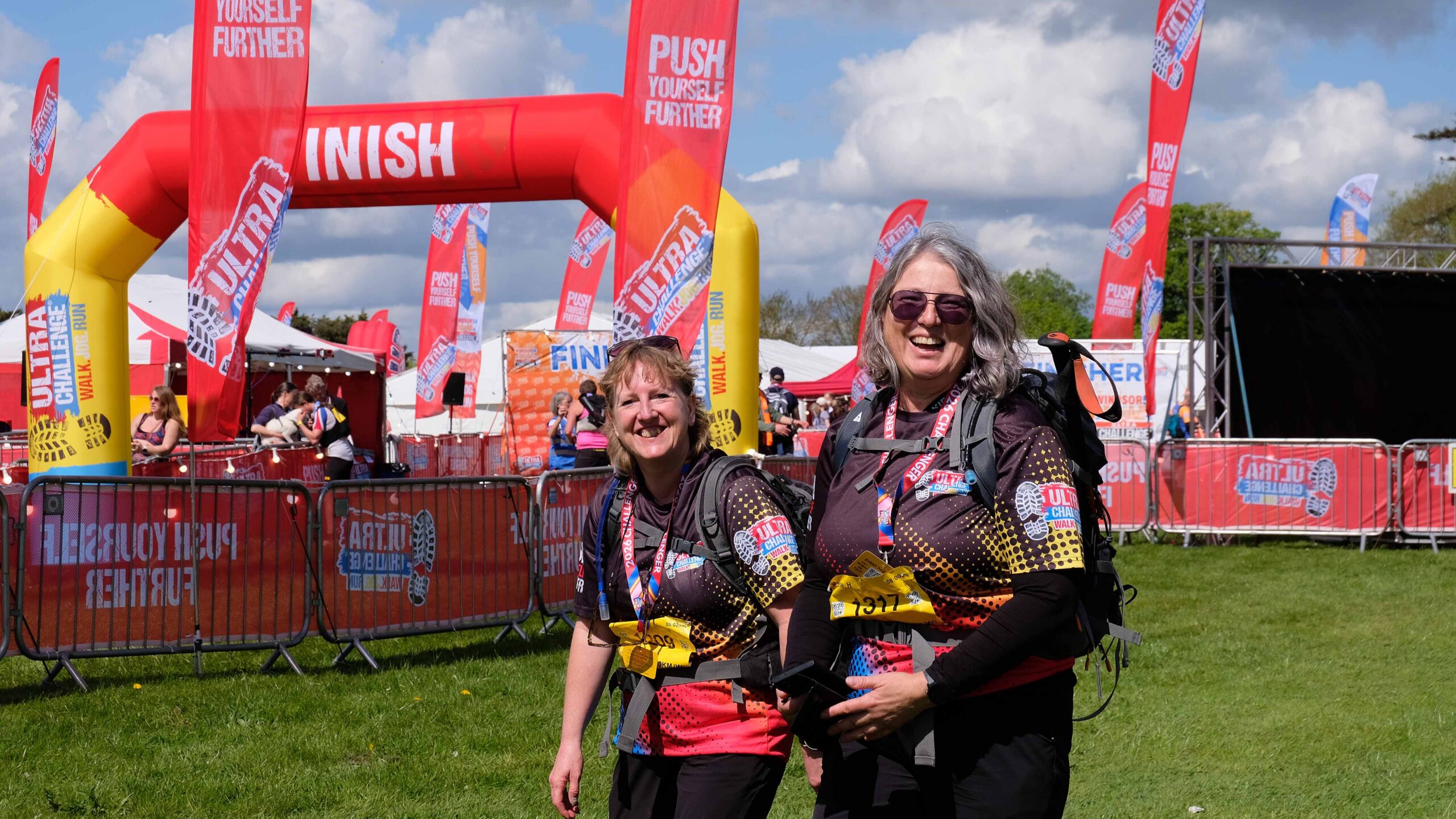 Two smiling women in colorful running gear pose at a lively charity race finish, with flags, barriers, crowds, and a red finish arch behind them.