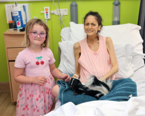 Alt text: A person sits in a hospital bed with a small skunk resting on a blue towel beside them. A child wearing glasses and a pink outfit stands next to the bed. The room includes medical equipment and a cabinet in the background. Text on the image reads: “A gift in your will can help families create precious memories together. This is hospice care.”