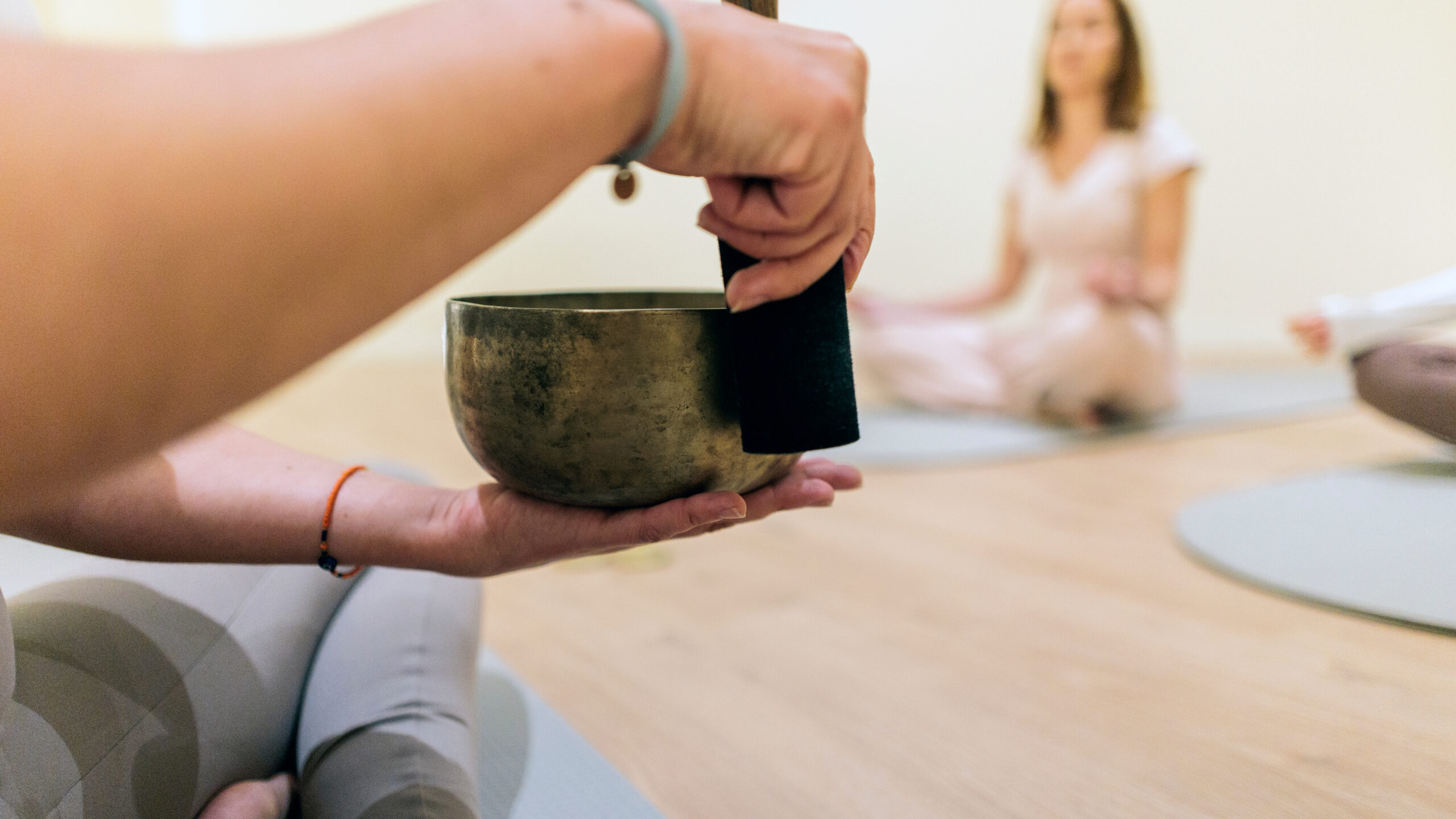 Person's hands gently playing an ancient singing bowl with a mallet, producing calming sounds during a collective sound bath meditation session focused on healing and relaxation